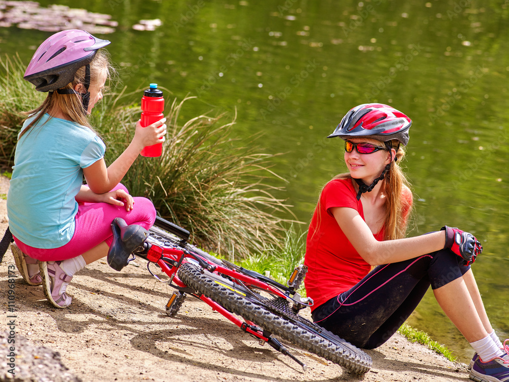 Obraz premium Bikes cycling family. Happy mother and daughter rides bicycle. Family have rest near water in park. Kid drinking bottle water. Bicycles are on coast. 