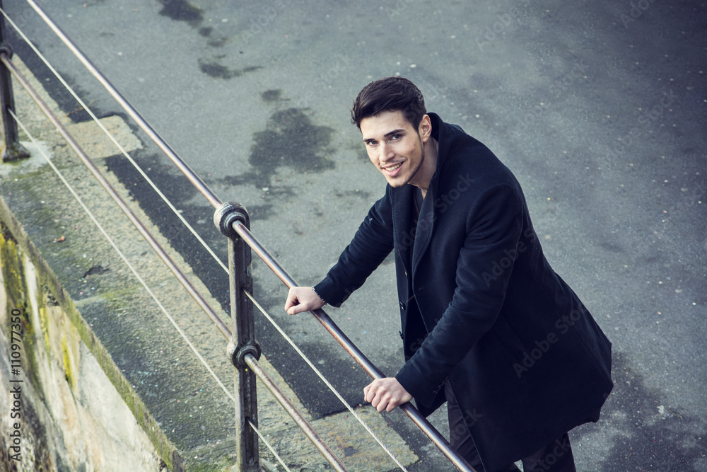 Young man in black coat looking down while leaning on railing