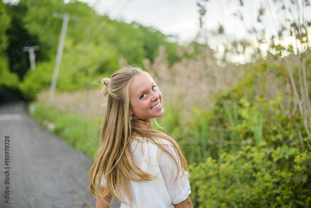 Portrait of young woman with long blond hair looking back on rural road Portrait of young woman with long blond hair looking back on rural road