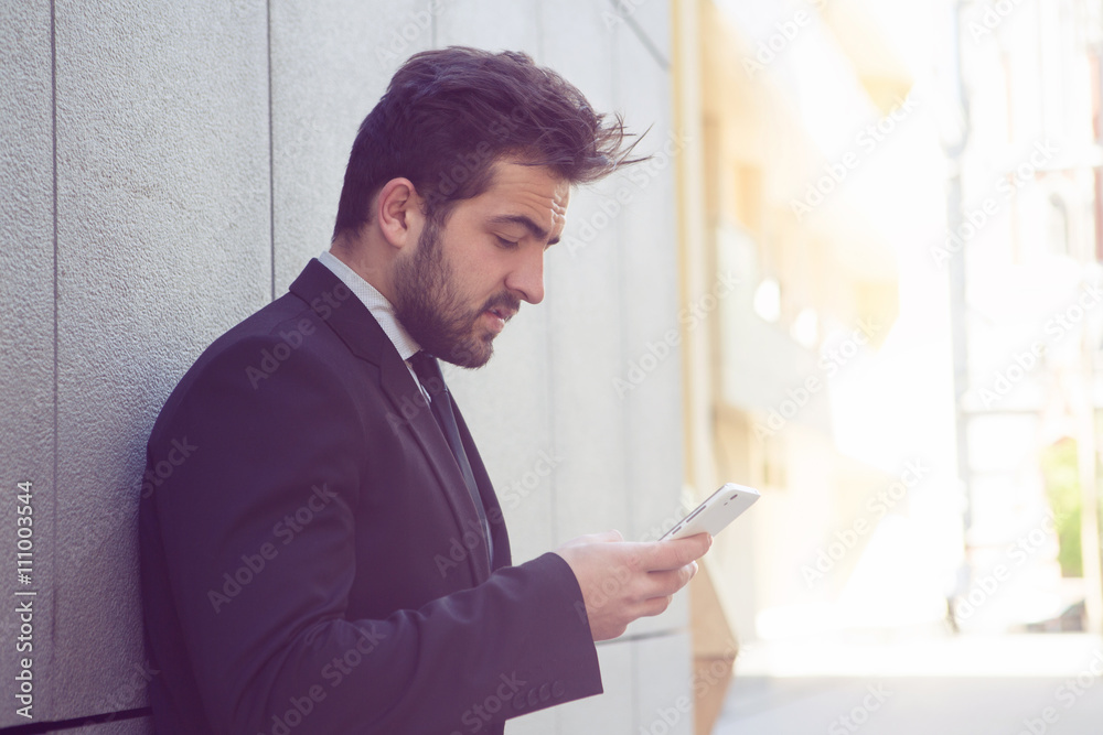 Toned picture of handsome businessman looking at mobile or smart phone's screen while stading near office building and waiting for someone.