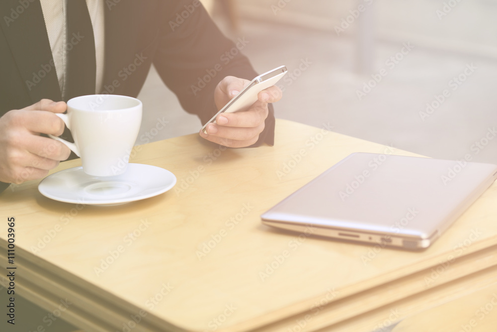 Closeup toned image of businessman using mobile phone while his laptop computer is represened in front of him. Freelance man drinking cup of tea or coffee. Closeup toned image of businessman using mobile phone while his laptop computer is represened in front of him. Freelance man drinking cup of tea or coffee.