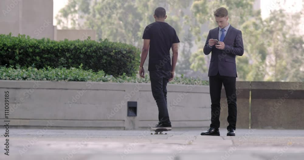 Smart young man in business suit texting on a cellphone on the streets in Downtown Los Angeles while a skateboarder passes in the foreground. Slow motion, wide shot recorded at 60fps. Smart young man in business suit texting on a cellphone on the streets in Downtown Los Angeles while a skateboarder passes in the foreground. Slow motion, wide shot recorded at 60fps.