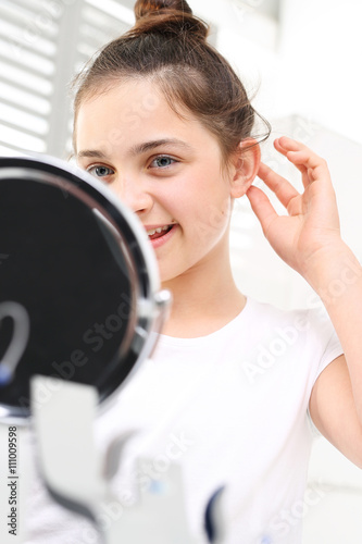 Aparat słuchowy dla dziecka. Wesoła dziewczynka zakłada aparat słuchowy przeglądając się w lusterku