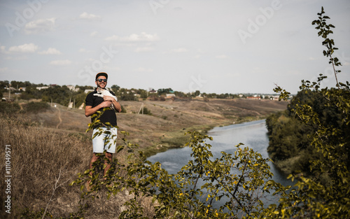 a bearded man in a black baseball cap, black shirt and shorts playing with white fluffy cat on top of a cliff on the background of the river and the forest