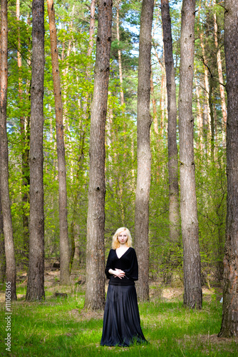 Beautiful smiling magic girl in the wood. Fashionable young short haired blond woman posing in the forest park wearing fancy empire style dress holding flowers. Concept of fantasy and magic.
