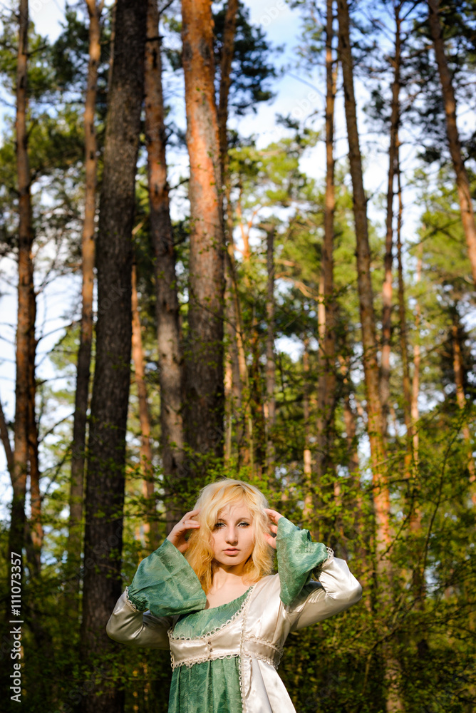 Fashionable shooting of a young short haired blond woman posing in the forest park wearing fancy empire style dress holding flowers. Concept of fantasy and magic. Fashionable shooting of a young short haired blond woman posing in the forest park wearing fancy empire style dress holding flowers. Concept of fantasy and magic.
