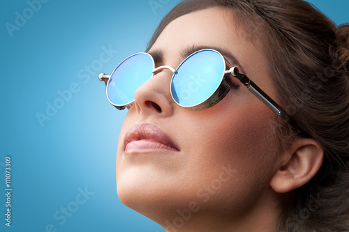 Close-up face portrait of young beautiful woman with perfect skin in round sunglasses looking up on blue background. Beauty face make-up.  © LazorPhotography