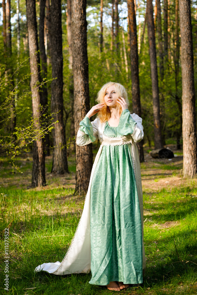  Fashionable shooting of a young short haired blond woman posing in the forest park wearing fancy empire style dress holding flowers. Concept of fantasy and magic.