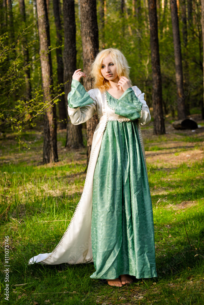  Fashionable shooting of a young short haired blond woman posing in the forest park wearing fancy empire style dress holding flowers. Concept of fantasy and magic.