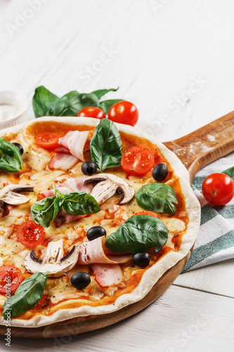 Close-up view of Italian PIZZA with mushrooms, basil, tomato, olives and cheese and bacon. WITHOUT one piece. white wooden table background. Look as Prosciutto, Capricciosa, Funghi, Cotto PIZZA. 