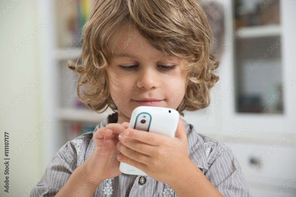 Cute little long haired boy happy sitting at the table and playing with smart phone. Enjoying modern generation technologies. Cute little long haired boy happy sitting at the table and playing with smart phone. Enjoying modern generation technologies.