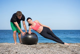 Sport und Fitness Instruktor zeigt junger Frau wie man mit Gymnastikball den Körper trainiert am Strand im Freien