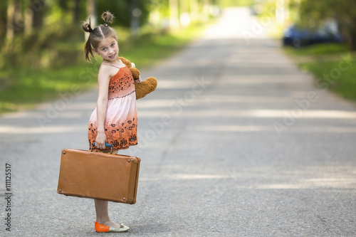 Cheerful little girl standing on the road with a suitcase and a Teddy bear.