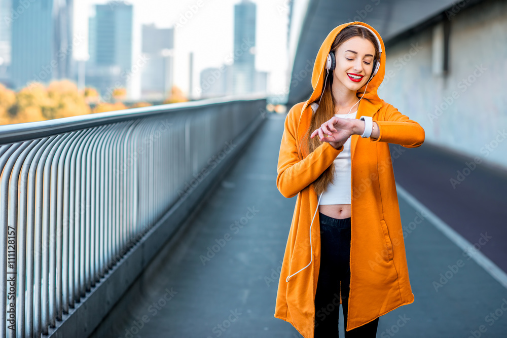 Fototapeta premium Young sport woman in yellow sweater listening to the music with smart watch on the modern bridge with skyscrapers on the background. Morning exercise in megacity