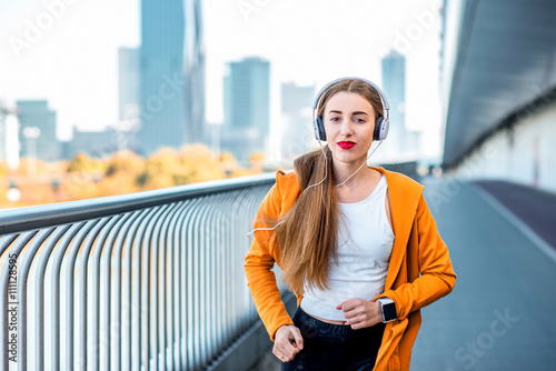 Photography Young sport woman in yellow sweater with earphones running on the modern bridge with skyscrapers on the background
