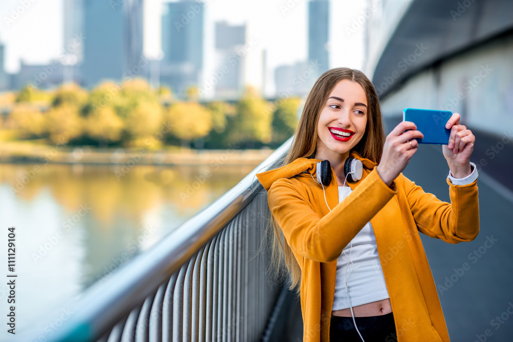 Fototapeta premium Young sport woman in yellow sweater making selfie photo with smart phone standing on the modern bridge with skyscrapers on the background.