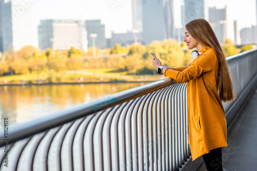Photography Young sport woman in yellow sweater using smart phone on the modern bridge with skyscrapers on the background