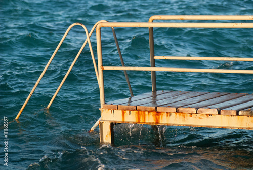 Old vintage metal wooden stairs leading to blue turquoise sea water from beach. Beautiful summer day. Swimming pool with grunge retro stairs on the ocean coast. Side View © skrotov