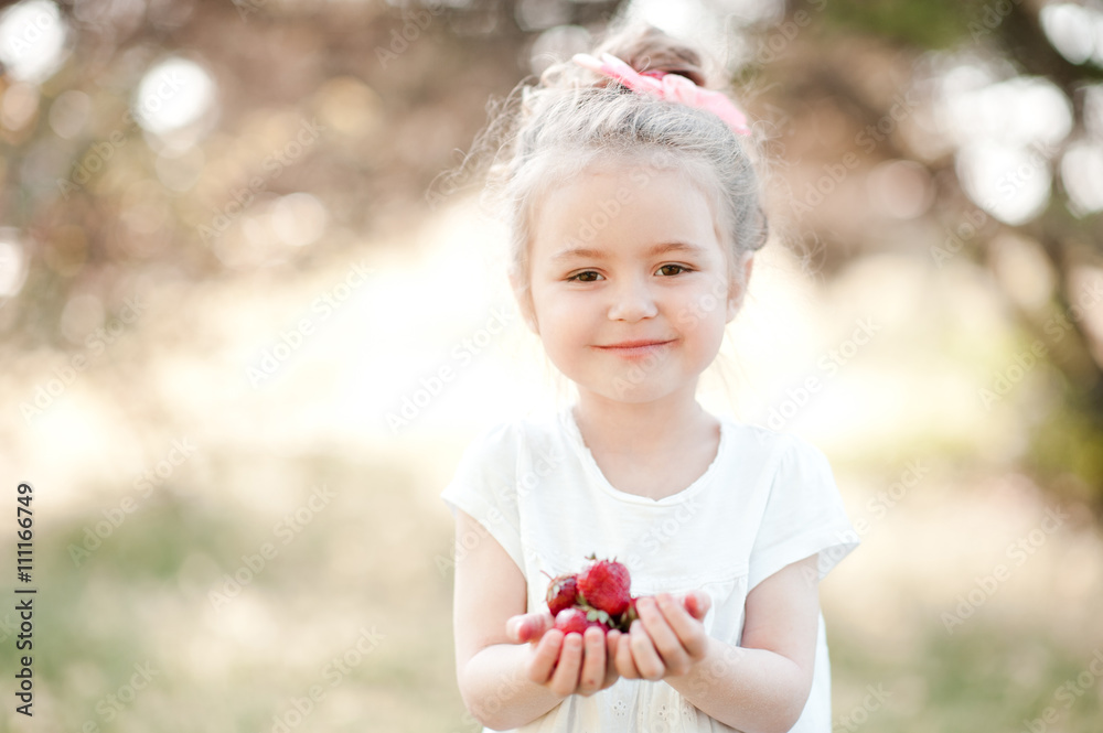 Cute kid girl 4-5 year old eating strawberries outdoors. Looking at camera. Childhood. Summer season.