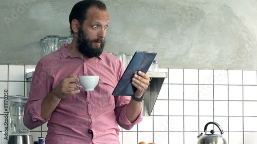 Young man reading shocking news on tablet computer and drinking coffee in kitchen at home 
