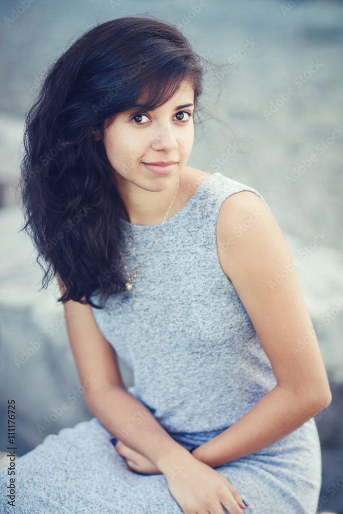 Portrait of beautiful Hispanic latino white girl woman with brown eyes, long dark curly wavy hair in gray dress sitting in park outside smiling laughing looking in camera, lifestyle portrait concept