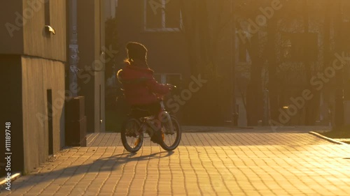 Kid is Riding a Bicycle Behind a Corner of House by City Street in the Evening Sunset Girl's Silhouette in the Sun Paving Stones Kid is Learning to Ride