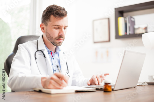 Young doctor taking notes and using a laptop