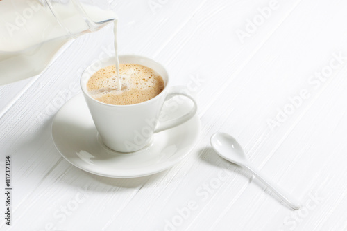 cup of hot cappuccino coffee on a white wood table. Milk being poured into a cup of coffee. Adding milk.