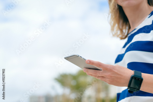 Side view of woman using smart watch with mobile phone on outdoors background. Close up photo
