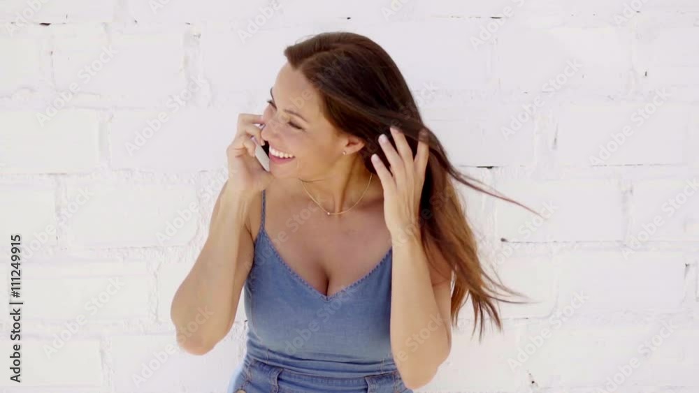 Smiling young woman talking on a mobile phone as she leans against a white brick wall in a trendy denim slack suit Smiling young woman talking on a mobile phone as she leans against a white brick wall in a trendy denim slack suit
