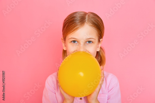Pretty little girl standing and inflating a yellow balloon on pink background