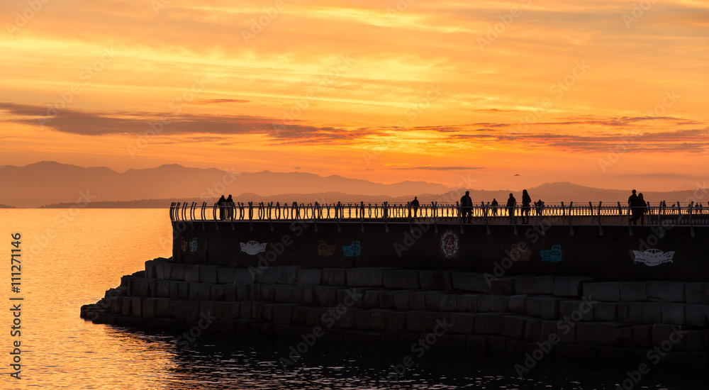Joggers and walkers are silhouetted by a beautiful sunset as they enjoy an evening on a breakwater at the harbor in Victoria Canada.