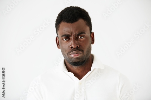 Close up shot of angry, grumpy or pissed off African American man with bad mood, looking and frowning at the camera, posing against white studio wall. Negative human face expressions and body language