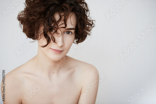 Cropped view of Caucasian female with perfect naked body looking and smiling at the camera with her brown curls falling down her face, posing against white copy space studio wall. Skin care concept