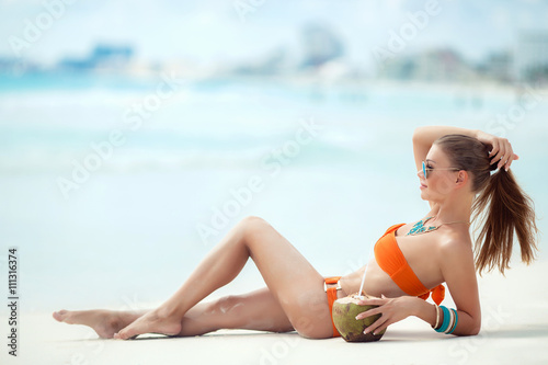 Young beautiful woman with long straight hair,sun glasses,slim figure,wears around his neck a large necklace,spends time on a tropical beach on white sand in orange bikini near the blue ocean