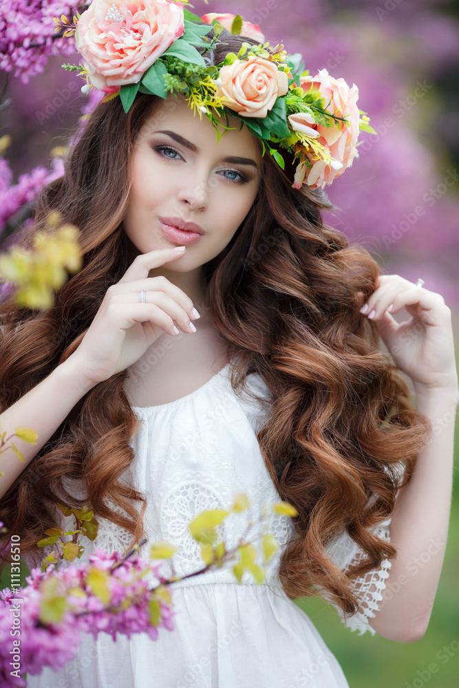 Spring portrait of a beautiful woman in a wreath of flowers,long curly hair red color,gray eyes,beautiful smile,wearing a white summer dress,posing near a bush blooming pink flowers outdoors in spring Spring portrait of a beautiful woman in a wreath of flowers,long curly hair red color,gray eyes,beautiful smile,wearing a white summer dress,posing near a bush blooming pink flowers outdoors in spring