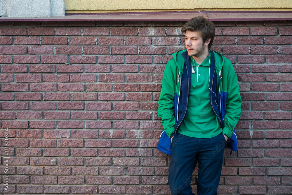 Common young man wearing green t-shirt jacket and jeans against a red brick wall looking sideways