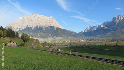 Idyllic scenery of Tyrolean countryside with railway tracks crossing green fields in the foreground and rugged Zugspitze Mountain in the background on a beautiful sunny day in Lermoos, Tirol, Austria