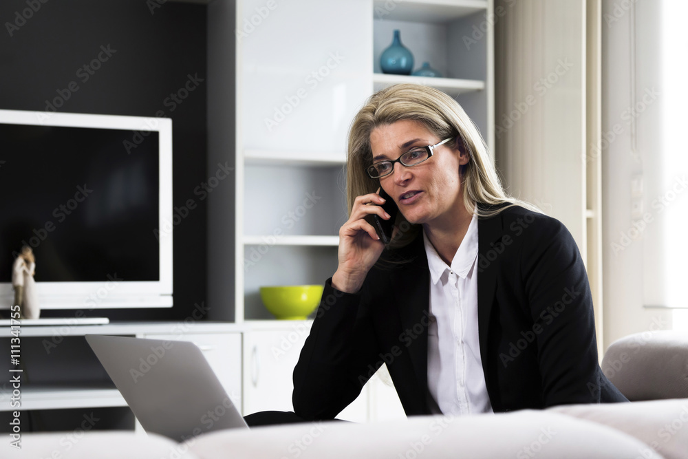Middle-aged and attractive business woman making a phone call while working on laptop computer. Modern home office in bright interior.