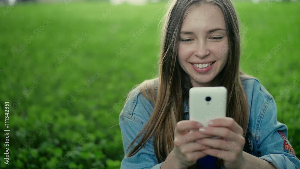 Young beautiful girl sitting on the grass and shows in the phone Young beautiful girl sitting on the grass and shows in the phone