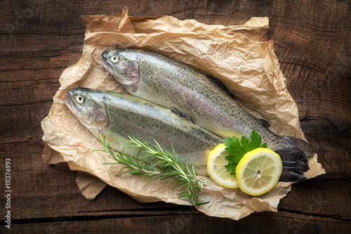 Fotografie Uncooked trout fish on parchment paper with rosemary and lemon against dark rust