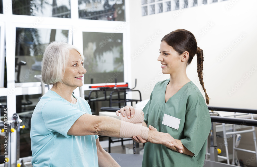 Smiling Female Nurse Putting Crepe Bandage On Senior Woman's Han Smiling Female Nurse Putting Crepe Bandage On Senior Woman's Han