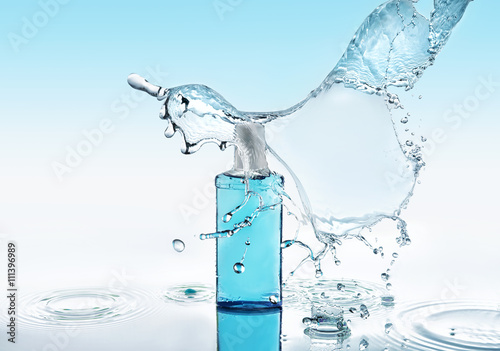 Blue moisturizing shampoo stays on the blue water background with big splash above the bottle and with many water drops and water waves around the bottle