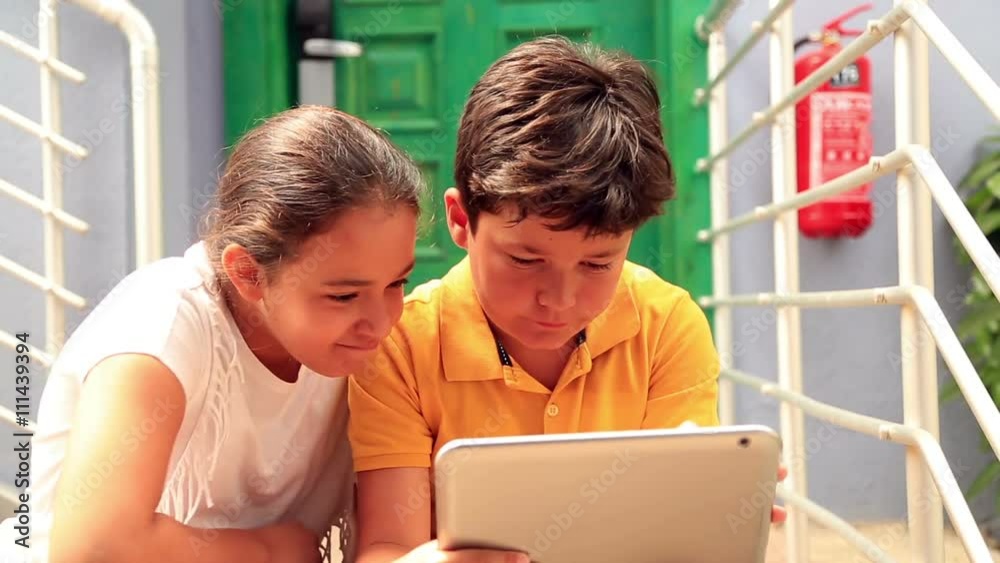 Closeup outdoor portrait of a cute kids in casual clothes sitting on the stairs in front of the door with digital tablet in her hands
