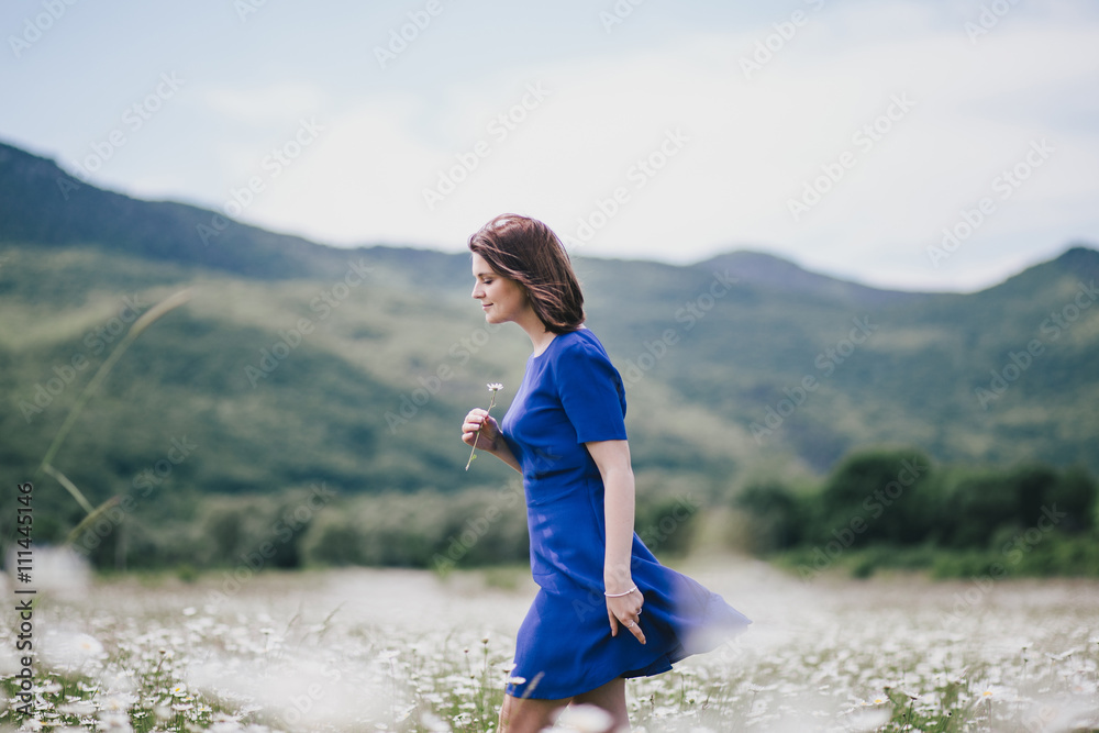 Young beautiful woman in a blue dress enjoying chamomile field among mountains. Summer mood Young beautiful woman in a blue dress enjoying chamomile field among mountains. Summer mood