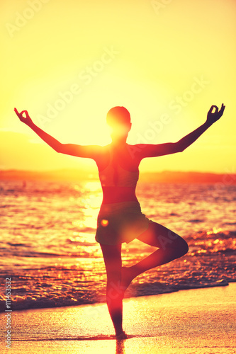 Fototapeta Naklejka Na Ścianę i Meble -  Wellness of mind - Yoga woman standing on one leg doing tree pose with open raised arms in sunset flare doing morning exercise routine on tropical beach. Mindfulness and meditation concept.