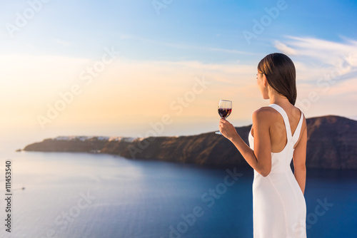 Young woman drinking red wine on outdoor terrace watching beautiful sunset view of Mediterranean Sea. Female in white sundress on summer Europe travel vacation in Santorini, Greece.