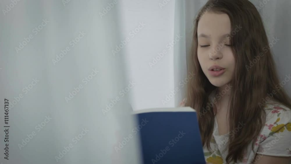 Beautiful little girl reading an interesting book by window.