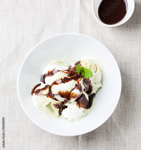 Vanilla and pistachio ice cream with chocolate sauce in a bowl on a plate on a linen textile, top view