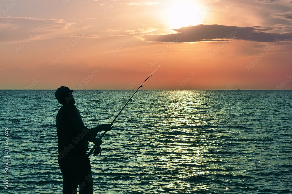 silhouette of a fisherman with a fishing rod in the sunset, dawn light casting a fishing rod from the sea shore. orange-red paint dawn. sun over the water.
